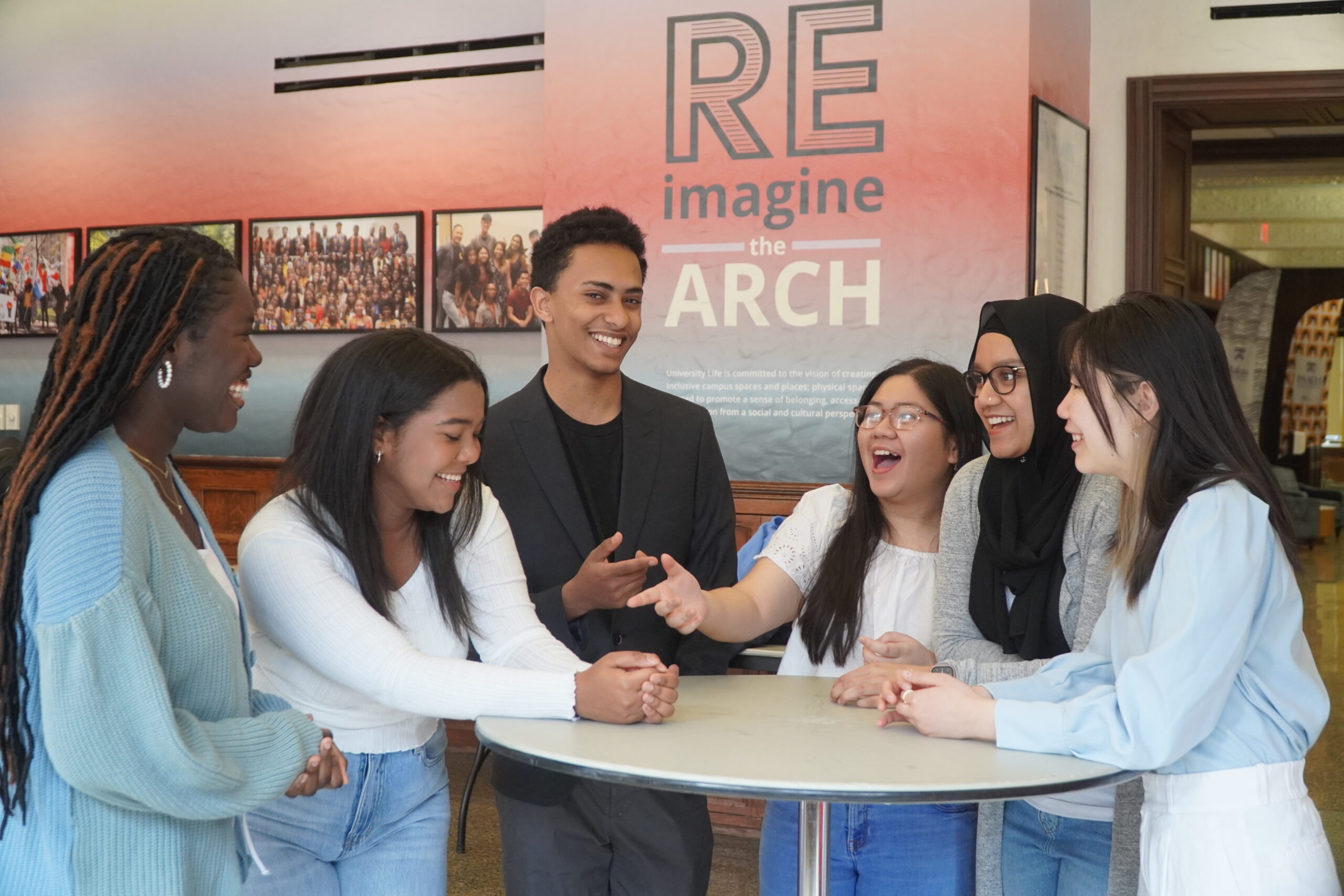Groups of six diverse students standing around a table laughing at a joke